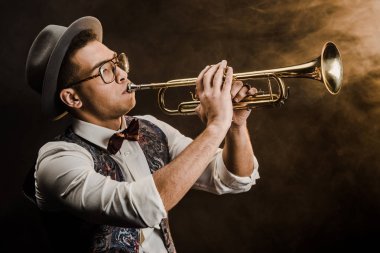 young mixed race jazzman in hat and eyeglasses playing on trumpet on stage with dramatic lighting and smoke clipart