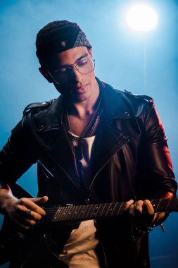 Erkek rock yıldızı deri ceketli elektro gitar üzerinde duman ve dramatik aydınlatma ile sahnede performans odaklı 