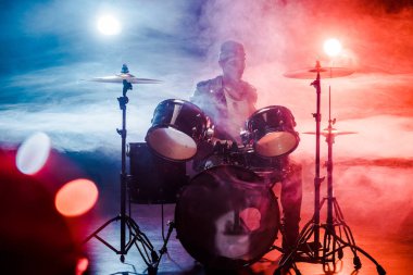 duman ve sahne ışıkları ile sahnede rock konseri sırasında davul çalmaya deri ceketli erkek müzisyen