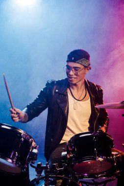 duman ve dramatik aydınlatma ile sahnede rock konseri sırasında davul çalmaya deri ceketli neşeli genç erkek müzisyen
