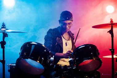 duman ve sahne ışıkları ile sahnede rock konseri sırasında davul çalmaya deri ceketli genç erkek müzisyen odaklı