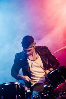 duman ve dramatik aydınlatma ile sahnede rock konseri sırasında davul çalmaya deri ceketli genç erkek müzisyen konsantre
