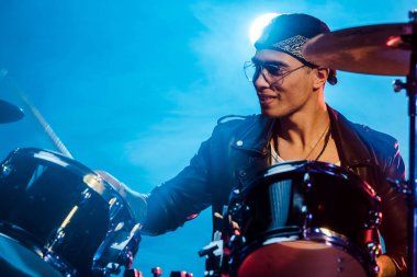 cheerful male musician in leather jacket playing drums during rock concert on stage with smoke and spotlight clipart