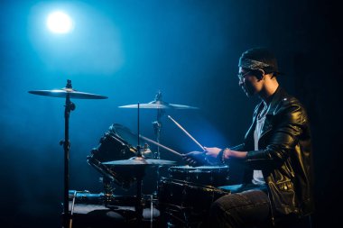 side view male musician in leather jacket playing drums during rock concert on stage with smoke and spotlight clipart