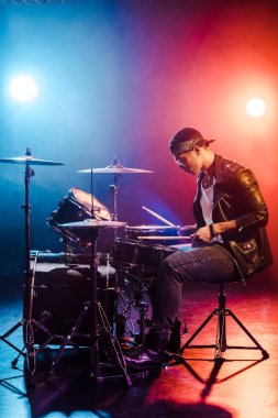 male rock star in leather jacket playing drums during concert on stage with smoke and spotlights clipart
