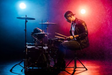 side view of male musician in leather jacket playing drums during rock concert on stage with smoke and spotlights