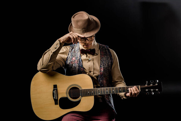 stylish mixed race male musician in eyeglasses and hat posing with acoustic guitar isolated on black