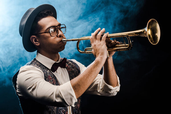 male musician in hat and eyeglasses playing on trumpet on stage with dramatic lighting and smoke
