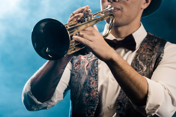 partial view of male musician playing on trumpet on stage with dramatic lighting and smoke