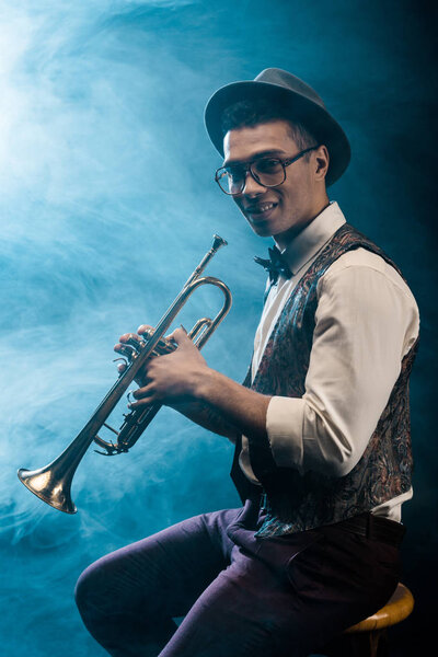 happy young jazzman posing with trumpet on stage with dramatic lighting and smoke