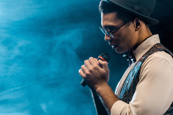 side view of handsome stylish man singing in microphone on stage with smoke and dramatic lighting 
