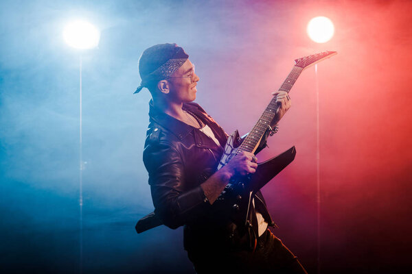 счастливый рок-звезда в кожаной куртке, выступающий на электрогитаре на сцене с дымом и драматическим освещением
 