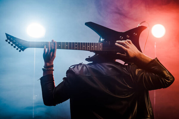 rear view of male musician in leather jacket playing on electric guitar behind head on stage
