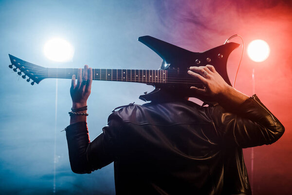 Задний вид мужчины-музыканта в кожаной куртке, играющего на электрогитаре за головой на сцене
