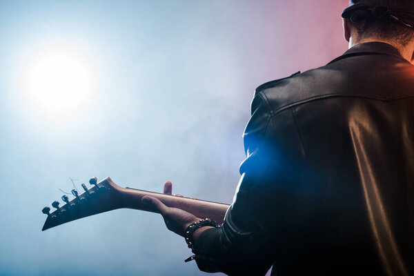 Задний вид рок-звезды в кожаной куртке, выступающей на электрогитаре на сцене с дымом и драматическим освещением
 