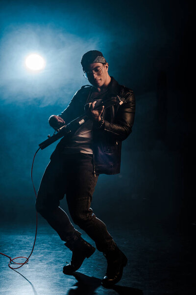 concentrated male rock star in leather jacket performing on electric guitar on stage with smoke and spotlight