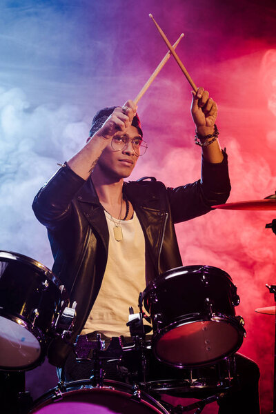 mixed race male musician raising drum sticks while sitting behind drum set on stage with dramatic lighting and smoke