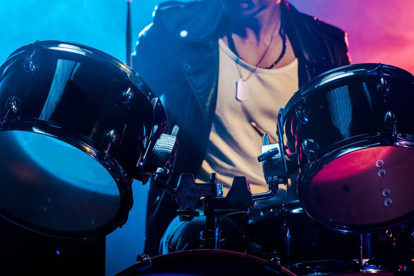 обрезанный кадр мужчины-музыканта, играющего на барабанах во время рок-концерта на сцене с дымом и драматическим освещением
