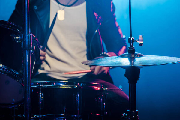 обрезанное изображение мужчины-музыканта, играющего на барабанах во время рок-концерта на сцене с дымом и драматическим освещением
