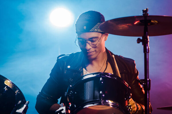 mixed race man in leather jacket playing drums during rock concert on stage with smoke and spotlight