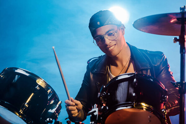 handsome mixed race male musician in leather jacket playing drums during rock concert on stage with smoke and spotlight