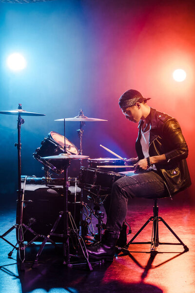 male rock star in leather jacket playing drums during concert on stage with smoke and spotlights