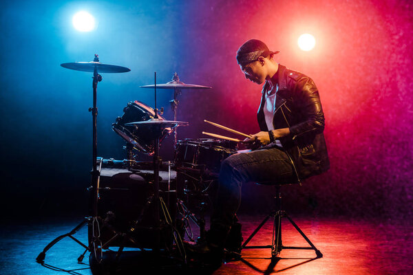 Вид сбоку мужчины-музыканта в кожаной куртке, играющего барабаны во время рок-концерта на сцене с дымом и прожекторами
