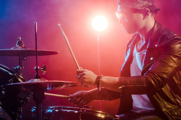 боковой вид молодого мужчины-музыканта в кожаной куртке, играющего барабаны во время рок-концерта на сцене
  