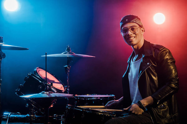 smiling mixed race male musician sitting behind drum set on stage with spotlights