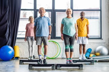 multiethnic senior athletes synchronous exercising on step platforms at gym clipart