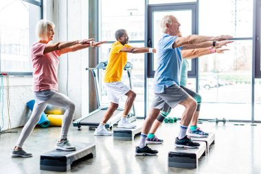 side view of multicultural senior athletes synchronous exercising on step platforms at gym clipart