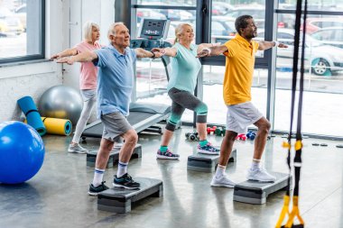 selective focus of senior multicultural athletes synchronous exercising on step platforms at gym clipart