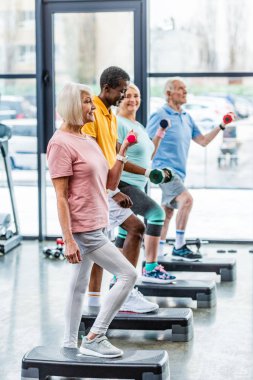 üst düzey çok kültürlü sporcu spor salonunda adım platformlarda dumbbells ile zaman uyumlu egzersiz yan görünüm