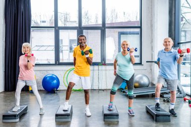 happy senior multicultural sportspeople synchronous exercising with dumbbells on step platforms at gym clipart
