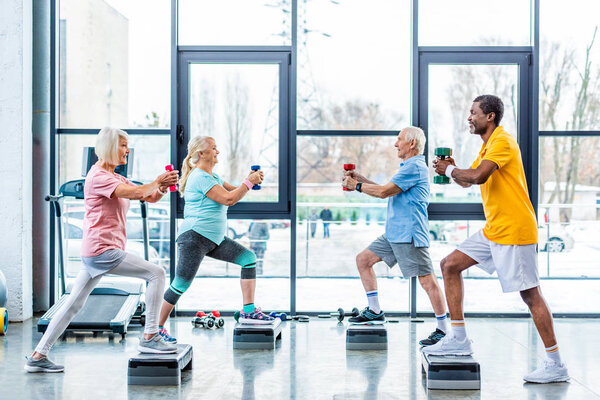 side view of senior multicultural sportspeople synchronous exercising with dumbbells on step platforms at gym