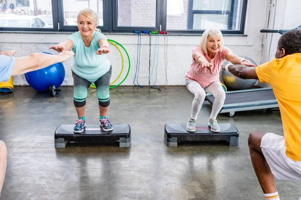Lächelnde Multiethnische Senioren Synchron Beim Kniebeugen Auf Trittbrettern Fitnessstudio — Stockfoto