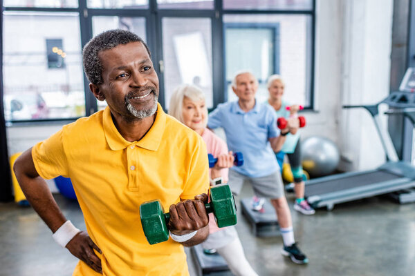 african american man and his friends synchronous exercising with dumbbells at sports hall