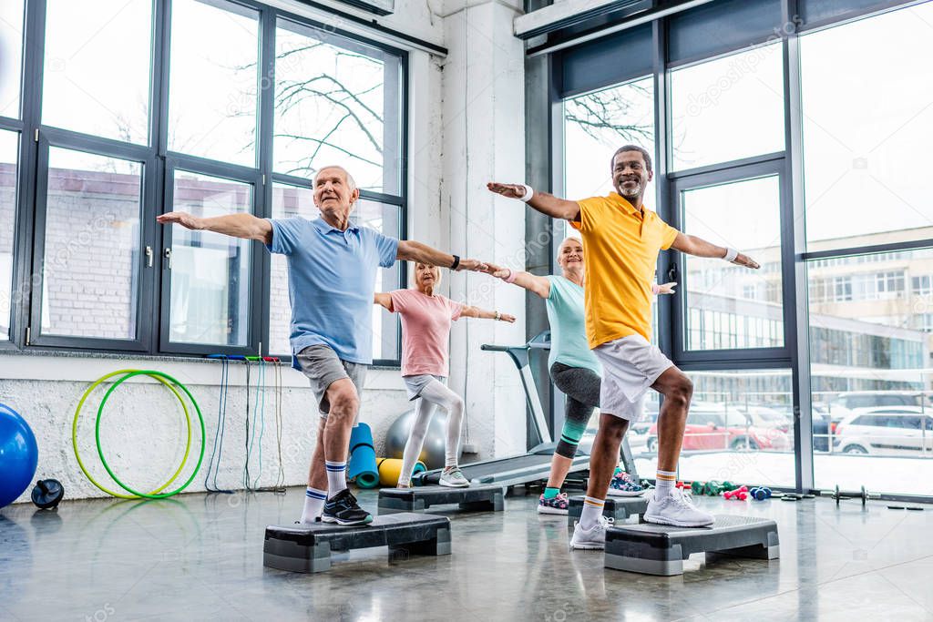 joyful senior multicultural athletes synchronous exercising on step platforms at gym