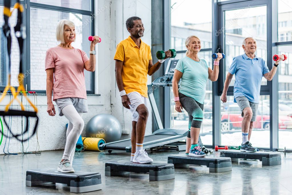 smiling senior multicultural sportspeople synchronous exercising with dumbbells on step platforms at gym