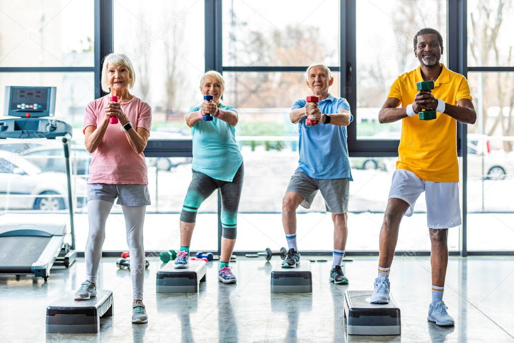 happy senior multicultural sportspeople synchronous exercising with dumbbells on step platforms at gym