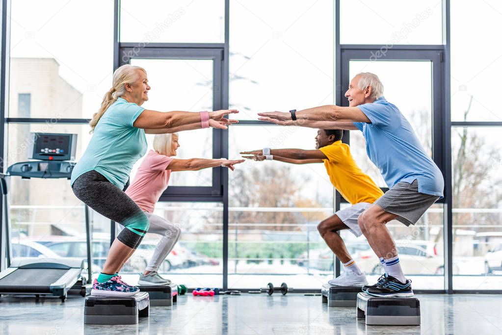 side view of multicultural senior athletes synchronous doing squats on step platforms at gym