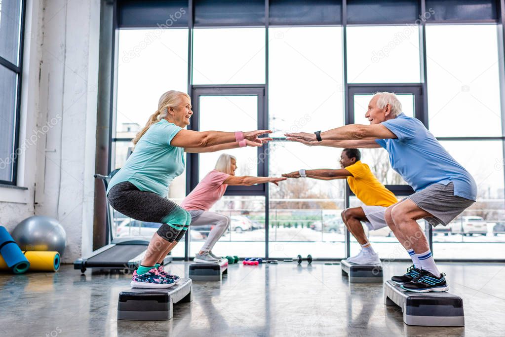 side view of senior multiethnic sportspeople synchronous doing squats on step platforms at gym