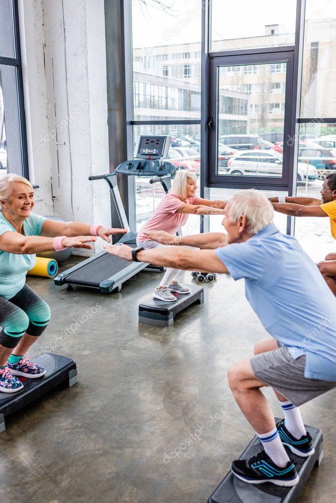 cheerful senior multiethnic sportspeople synchronous doing squats on step platforms at gym