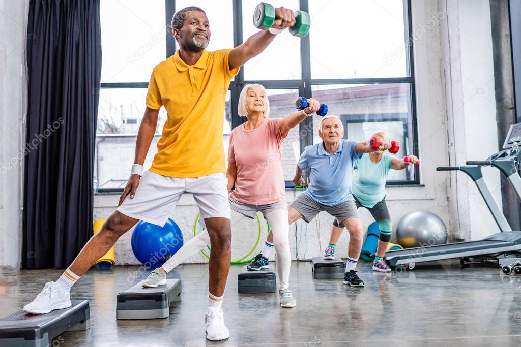 senior sportspeople synchronous exercising with dumbbells on step platforms at gym