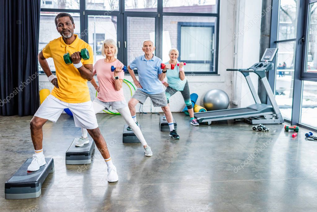 multiethnic senior sportspeople synchronous exercising with dumbbells on step platforms at gym