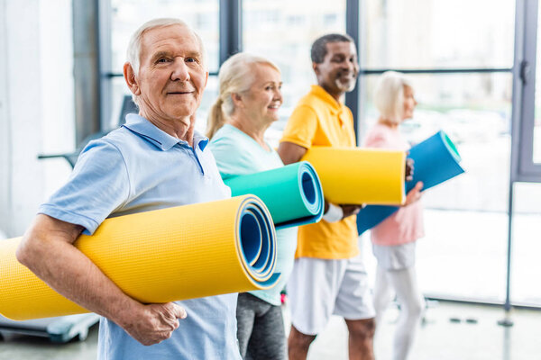 selective focus of senior sportsman holding fitness mat and his friends standing behind at gym