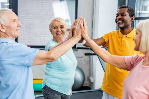 smiling multicultural senior athletes stacking hands together at gym