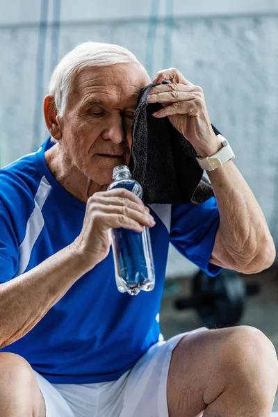 水のボトルを押しながらジムで休んでタオルで頭を拭く疲れてシニアのスポーツマン — ストック写真