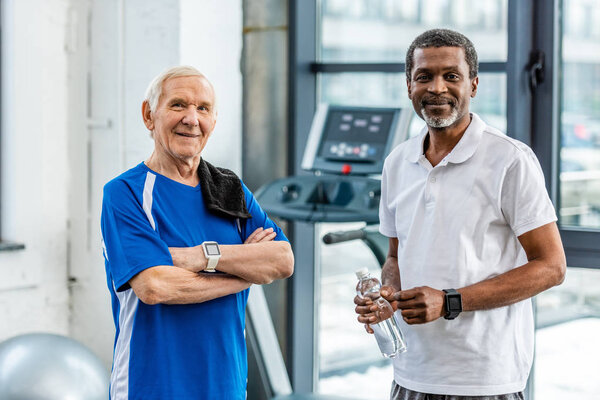 happy multicultural mature sportsmen with smartwatches standing at gym