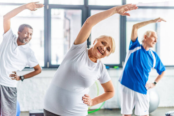 selective focus of smiling senior sportswoman synchronous exercising with male friends at gym
